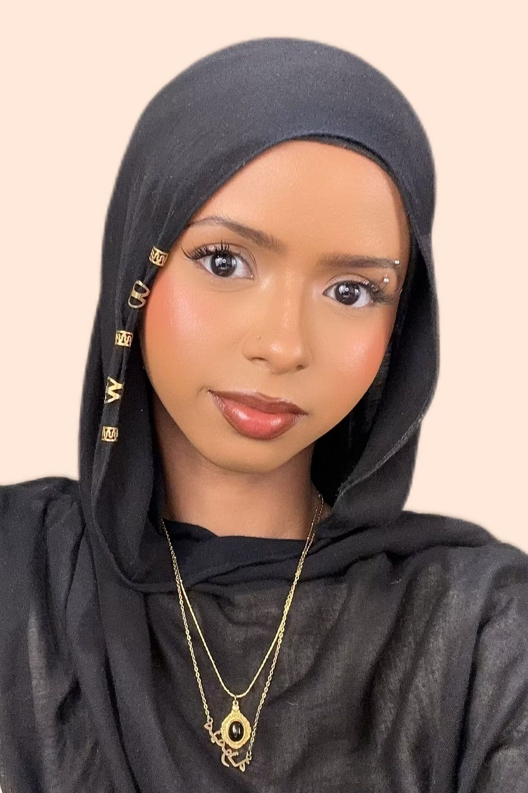 Woman wearing a black hijab and gold necklace on a beige background